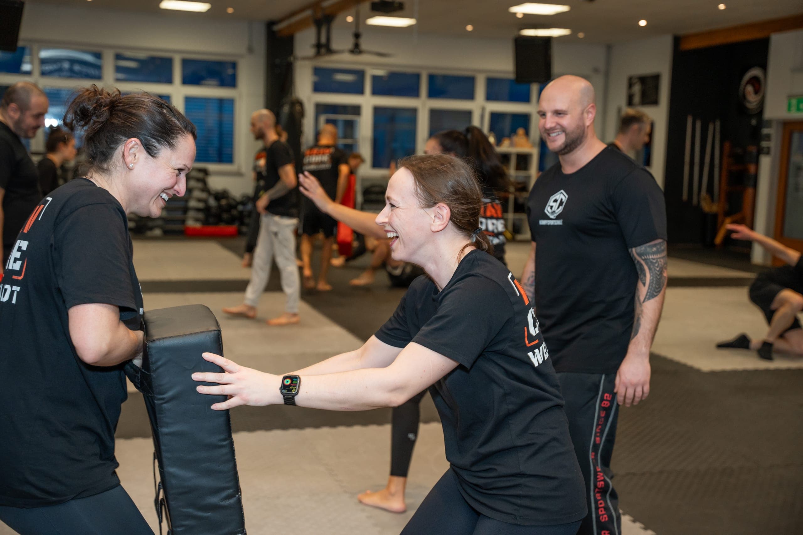Zwei Erwachsene lachen während einer Partnerübung an der Pratze, der Trainer steht daneben