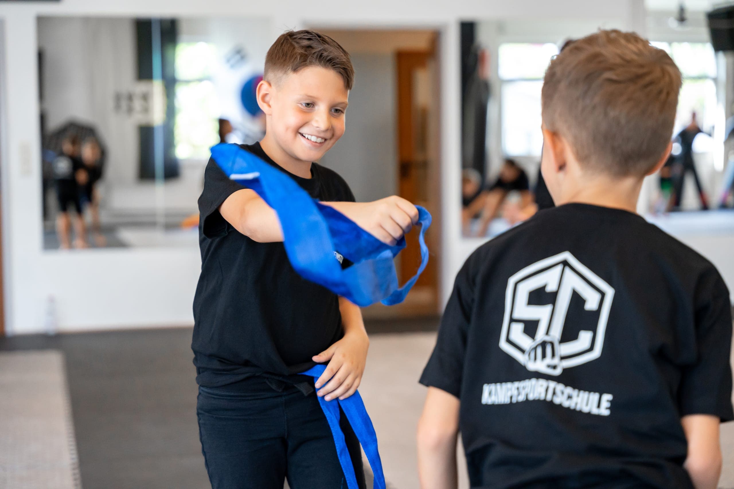 Zwei Kinder binden im Training ihren blauen Gürtel und lachen dabei